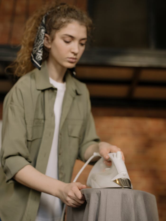 A young woman with curly hair ironing clothes in a cozy home interior with brick wall background.